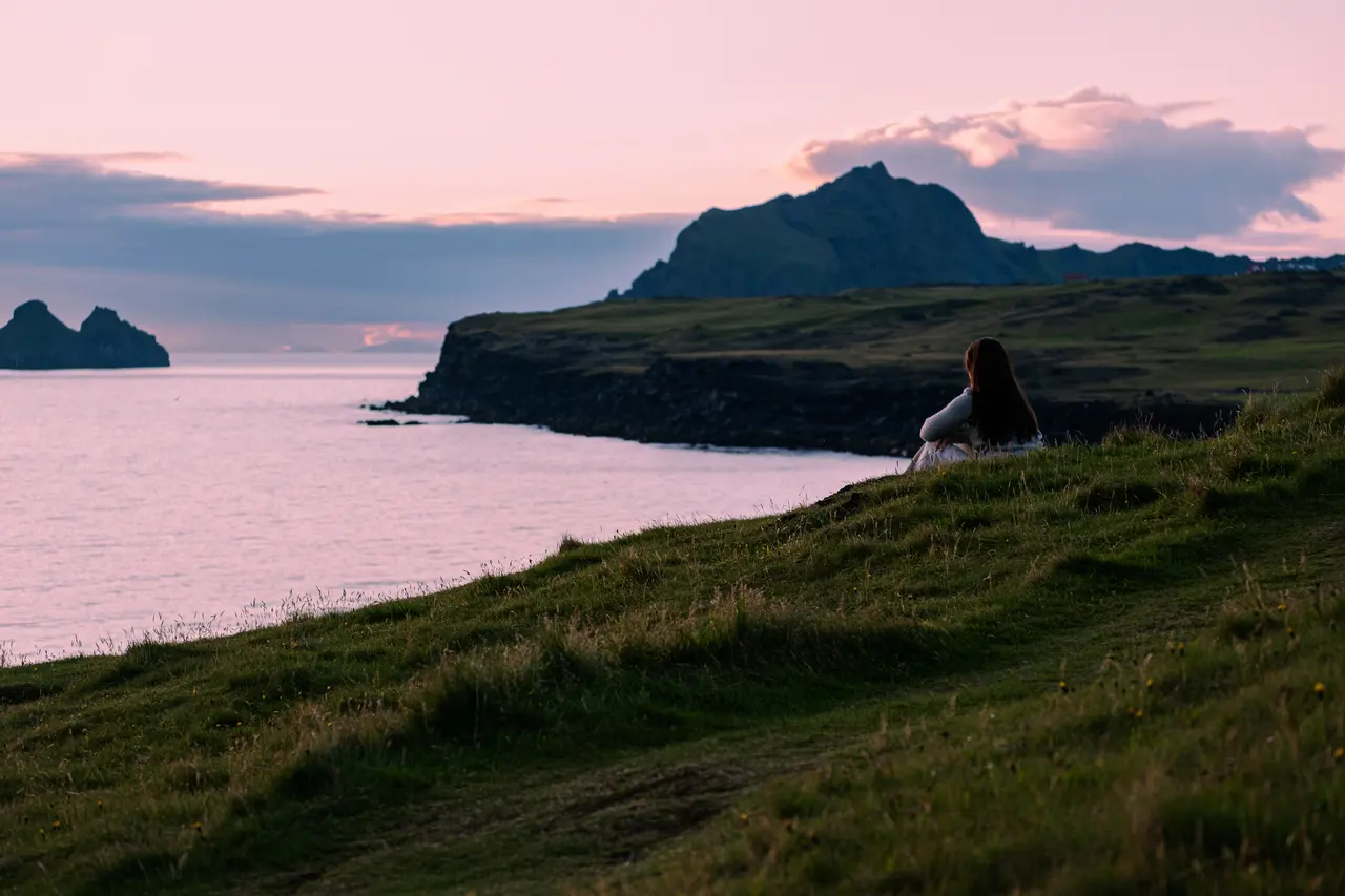 A woman sitting on a grass headland at dusk above flat pink water