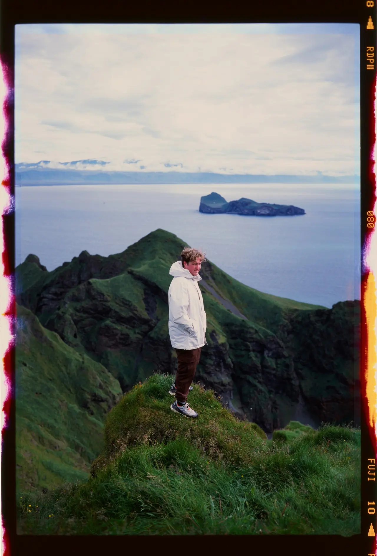 A figure on a grassy peak above the sea, an offshore island and the snow-streaked mainland behind