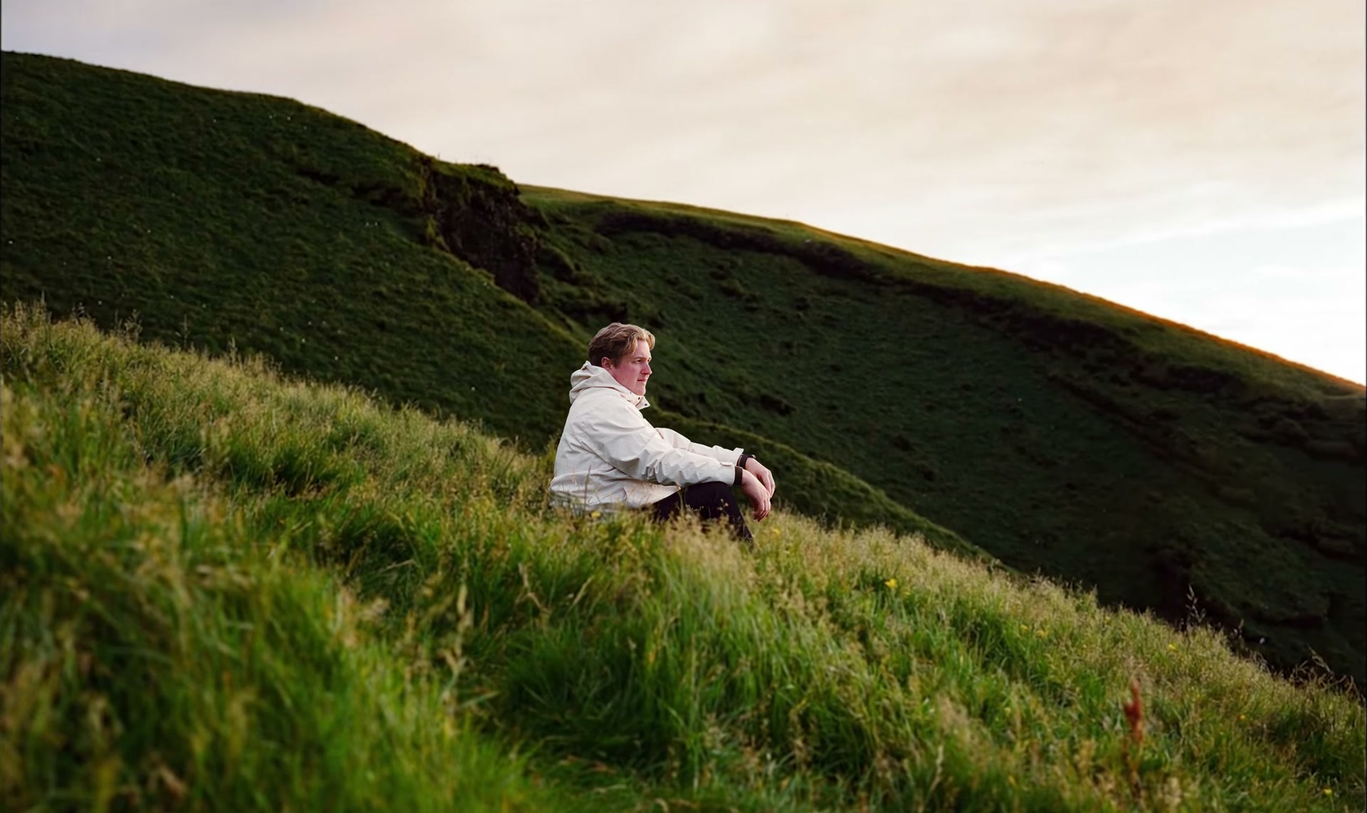 A person sitting in long grass on a green Icelandic hillside, looking out along the ridge as the light goes.